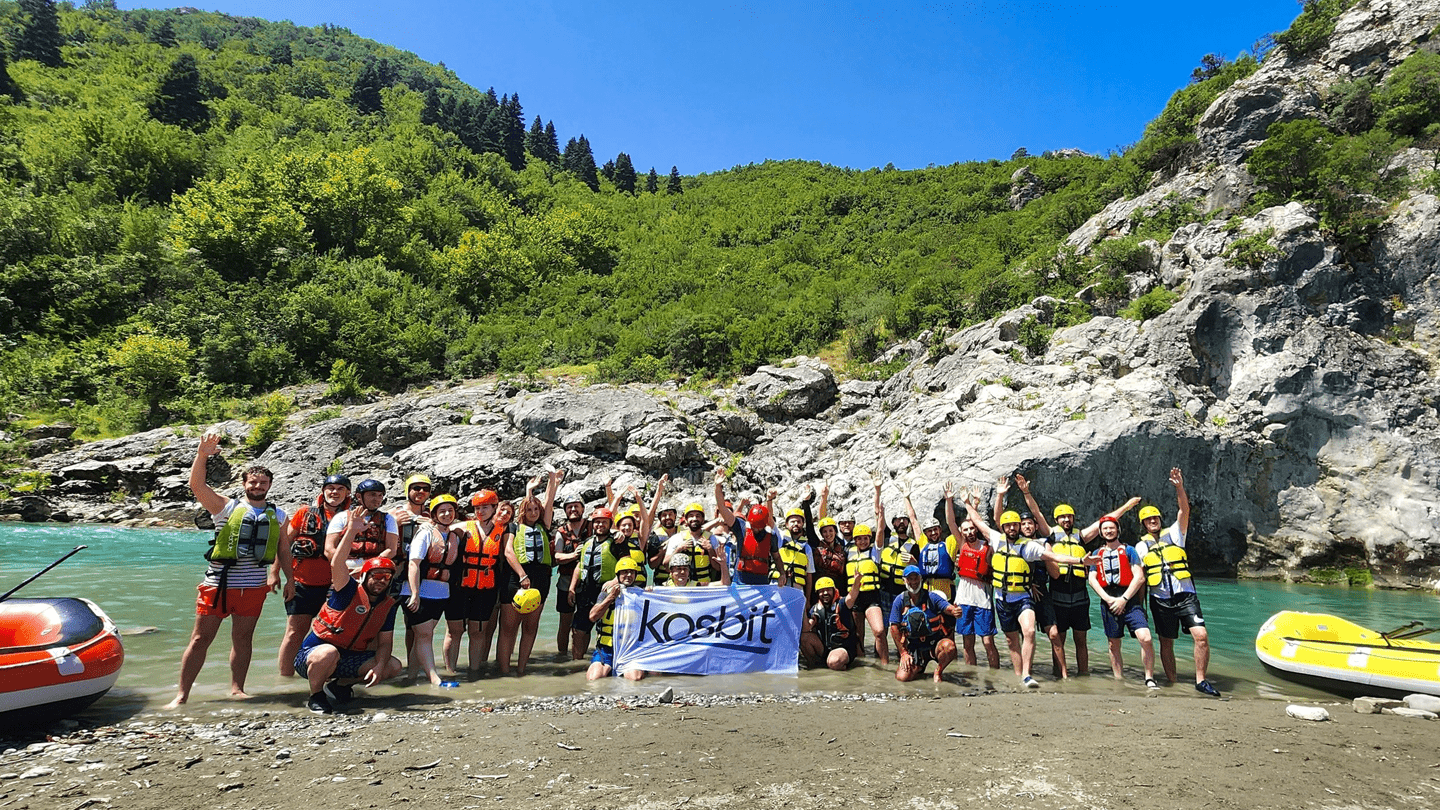 Group of Kosbit employees in life vests posing by the river during a rafting team-building retreat, surrounded by green hills and clear blue water.
