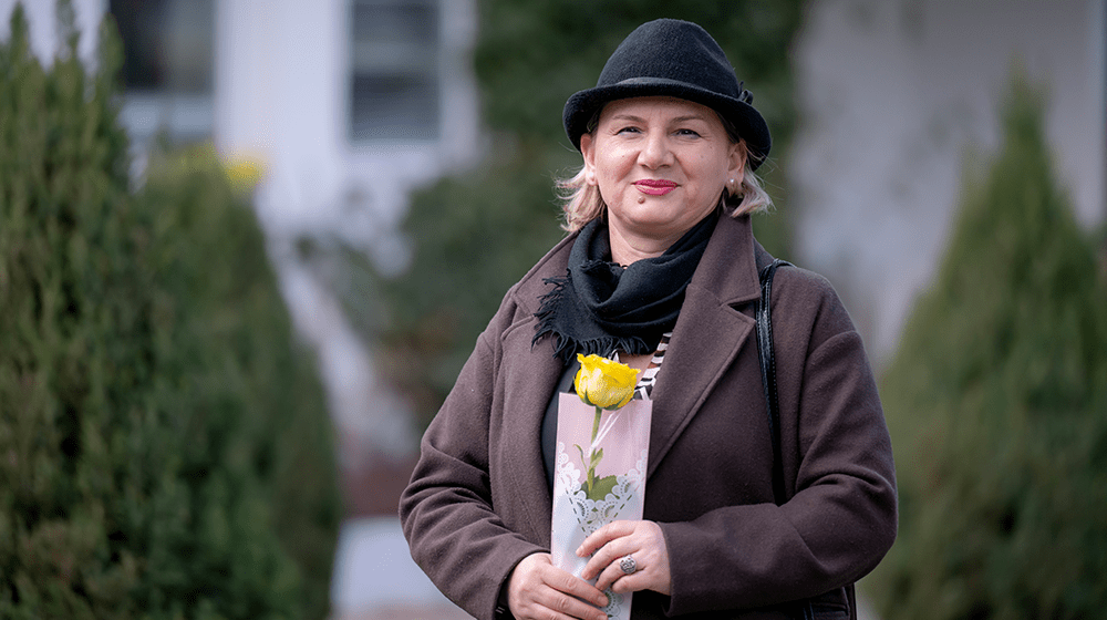 A woman with a black hat and a brown coat posing for a picture holding a yellow flower